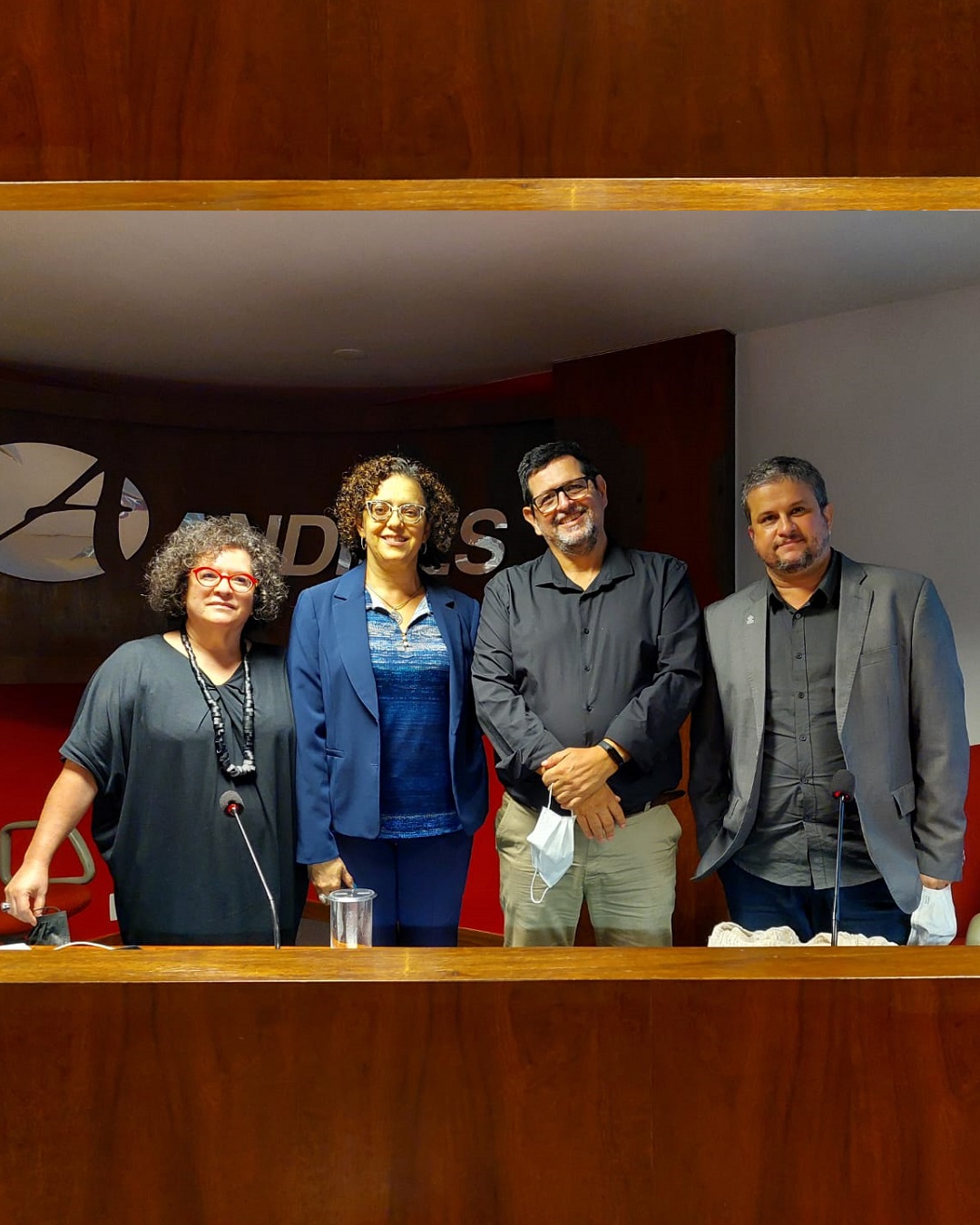 Descrição da imagem: fotografia colorida. Quatro pessoas encontram-se posicionadas atrás  de uma mesa, uma ao lado da outras. Aparecem da cintura para cima. Da direita para a esquerda seguem dois homens e duas mulheres. Todos estão de frente para o expectador. Ao fundo uma parede branca com um painel preto onde se lê: Andifes.