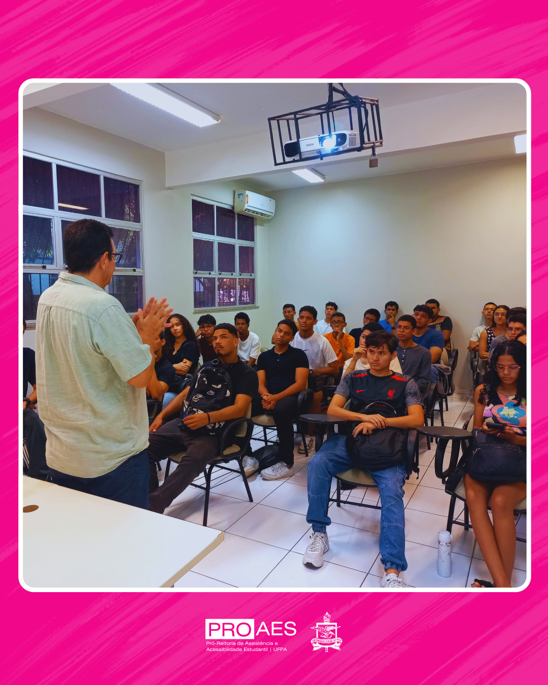 O card apresenta uma fotografia centralizada, com moldura branca e fundo rosa vibrante, com texturas diagonais mais claras. Na parte inferior do card, sobre o fundo rosa, há dois logotipos: à esquerda, o da "PROAES" (Pró-Reitoria de Assistência e Acessibilidade Estudantil da UFPA) em letras maiúsculas brancas; à direita, o brasão oficial da UFPA, também em branco.

Descrição da fotografia:

Em uma sala de aula bem iluminada, com paredes claras e piso branco, cerca de trinta estudantes estão sentados em cadeiras universitárias, prestando atenção a um homem que está de pé, de costas para a câmera, aparentemente conduzindo uma fala ou apresentação. Ele veste uma camisa verde-clara e óculos, gesticulando com as mãos. Entre os estudantes, há diversidade de gênero e estilo; alguns seguram mochilas ou garrafas d’água, enquanto outros observam atentamente ou manuseiam o celular.

No teto, destaca-se um projetor branco, protegido por uma estrutura metálica. O ambiente possui janelas amplas com grades, por onde é possível ver uma vegetação externa. O ar-condicionado está instalado acima de uma das janelas, sugerindo um espaço climatizado e confortável.

Elementos gráficos:

Moldura arredondada branca ao redor da fotografia.

Fundo rosa-choque com textura de linhas inclinadas.

Logos institucionais em branco na base do card.

Esse card transmite uma atmosfera institucional, acolhedora e informativa, com foco na participação estudantil e nas ações da PROAES/UFPA.